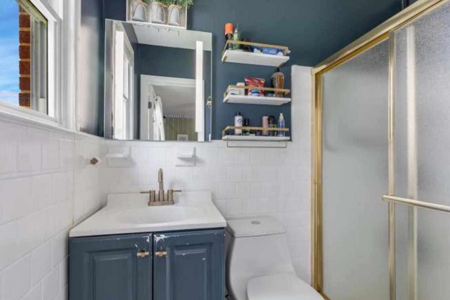 A bathroom with a white sink, a toilet, and a shower area. There are shelves above the toilet with toiletries.