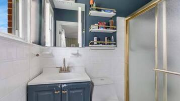 A bathroom with a white sink, a toilet, and a shower area. There are shelves above the toilet with toiletries.