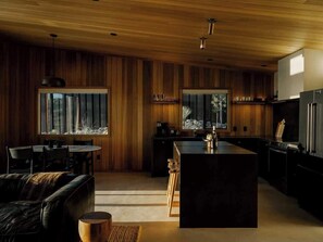 A modern kitchen with wooden paneling, a central island, and a dining area with a sofa and chairs. - Secluded Retreat | Two Homes, Pool + Firepit (Yucca Valley)
