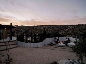 A modern outdoor seating area with a fire pit, surrounded by a white fence, overlooking a desert landscape. - Secluded Retreat | Two Homes, Pool + Firepit (Yucca Valley)