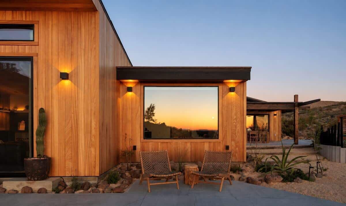 A modern wooden house with large windows, outdoor seating, and a desert landscape view.