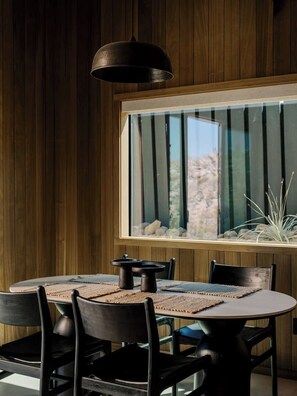 A dining area with a round table, black chairs, and a large window with a view of rocks and plants. - Secluded Retreat | Two Homes, Pool + Firepit (Yucca Valley)