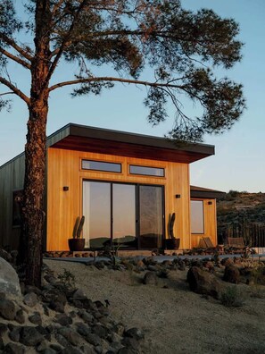 A modern building with a wooden facade, large sliding glass doors, and a desert landscape with rocks and sparse vegetation. - Secluded Retreat | Two Homes, Pool + Firepit (Yucca Valley)