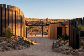 A modern building with a wooden facade, a stone pathway, and a desert landscape in the background. - Secluded Retreat | Two Homes, Pool + Firepit (Yucca Valley)
