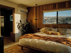 A bedroom with a large bed, a view of a desert landscape, a potted plant, and a wooden wall panel. - Secluded Retreat | Two Homes, Pool + Firepit (Yucca Valley)
