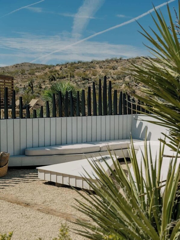 A modern outdoor seating area with a white cushioned bench and a wooden fence. - Secluded Retreat | Two Homes, Pool + Firepit (Yucca Valley)