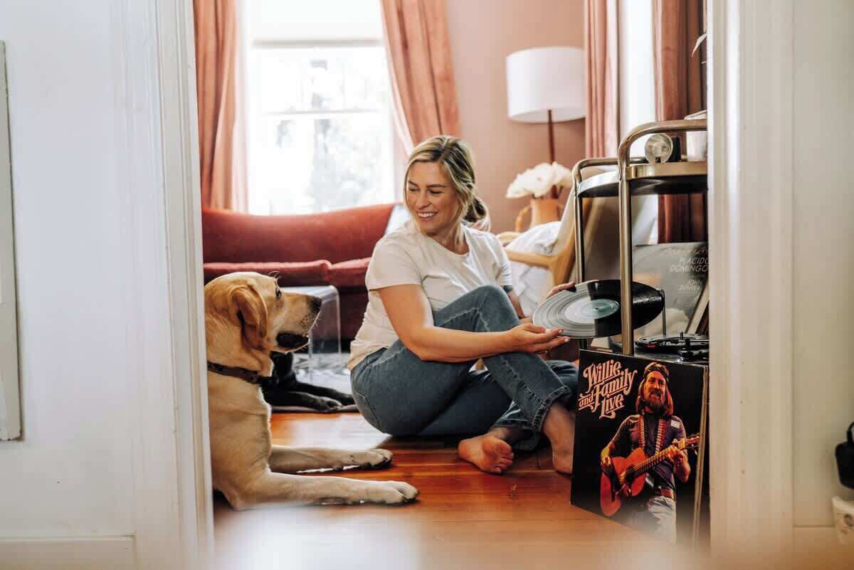 En person sidder på gulvet med en hund, mens vedkommende holder i en vinylplade, i et rum med en rød sofa og en lampe i baggrunden.