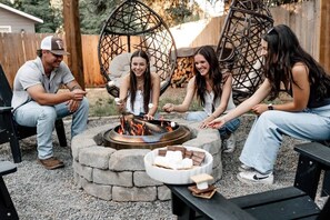 En gruppe mennesker sidder på udendørs møbler omkring et bålsted med marshmallows.