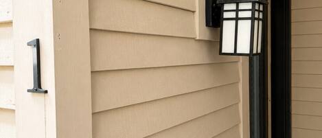 Pared exterior color beige con revestimiento horizontal, un marco de puerta negro y un accesorio de luz negro fijado a la pared.