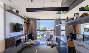 Quarto de hotel moderno com uma janela grande, uma televisão de tela plana, um espaço de jantar com mesa posta para dois e uma área de estar com sofá.