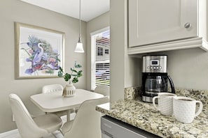 A kitchen with a coffee maker, two mugs, a table with chairs, a hanging light, and a framed floral artwork. - Island Living at Isla Del Sol | Ground Floor 2BR/2BA Getaway (Isla Del Sol)