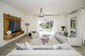 A spacious living room with a large TV, a wooden shelf, a sofa, a coffee table, and a view of a pool through the glass doors. - Private 4BR Villa with Pool inside All-Inclusive Beach Resort, Puerto Plata (Puerto Plata)