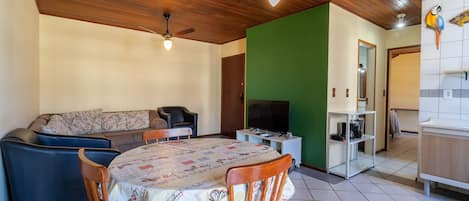 A dining area with a table and chairs, a sofa, a television, and a kitchen area with cabinets and a refrigerator.