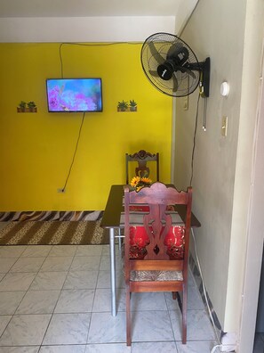 Ein Zimmer mit gelber Wand, einem daran befestigten Fernseher, einem Standventilator, einem Holzstuhl mit Kissen und einem gemusterten Teppich auf dem Boden.