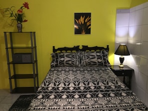 Un letto con una coperta a motivi, una lampada da comodino, uno scaffale con un vaso di fiori e un quadro appeso al muro.