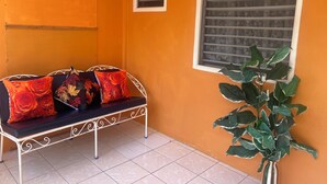 Quarto com um banco de metal com almofadas, uma planta em vaso e uma janela com persianas.