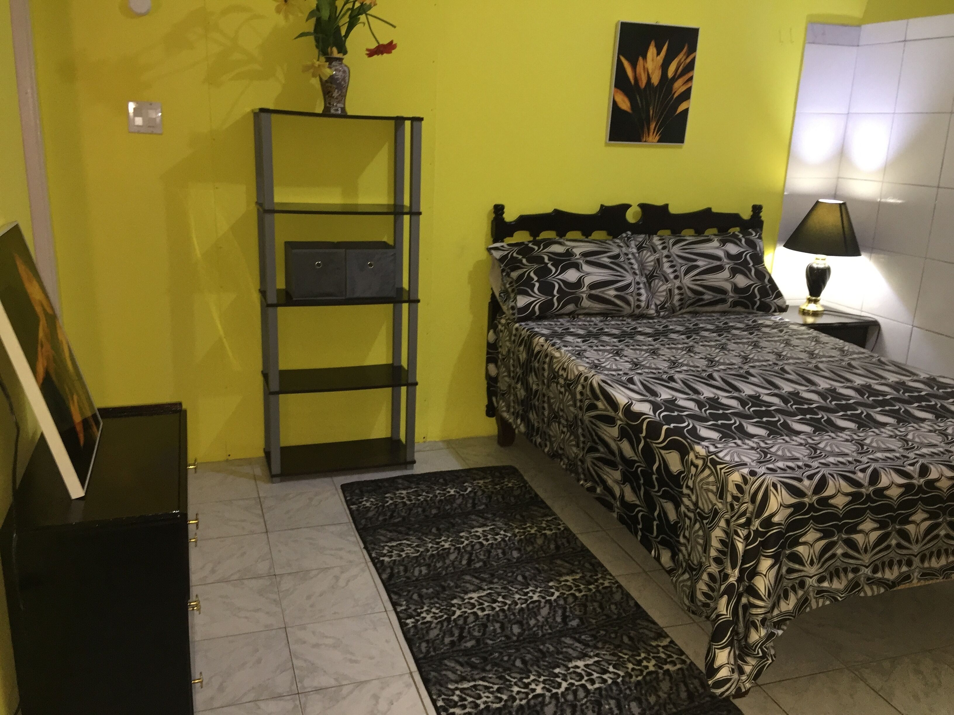 Ein Schlafzimmer mit einem Bett, einem Nachttisch, einer Lampe, einem Regal mit Aufbewahrungsboxen, einer Vase mit Blumen und einem gerahmten Bild auf einem Ständer.