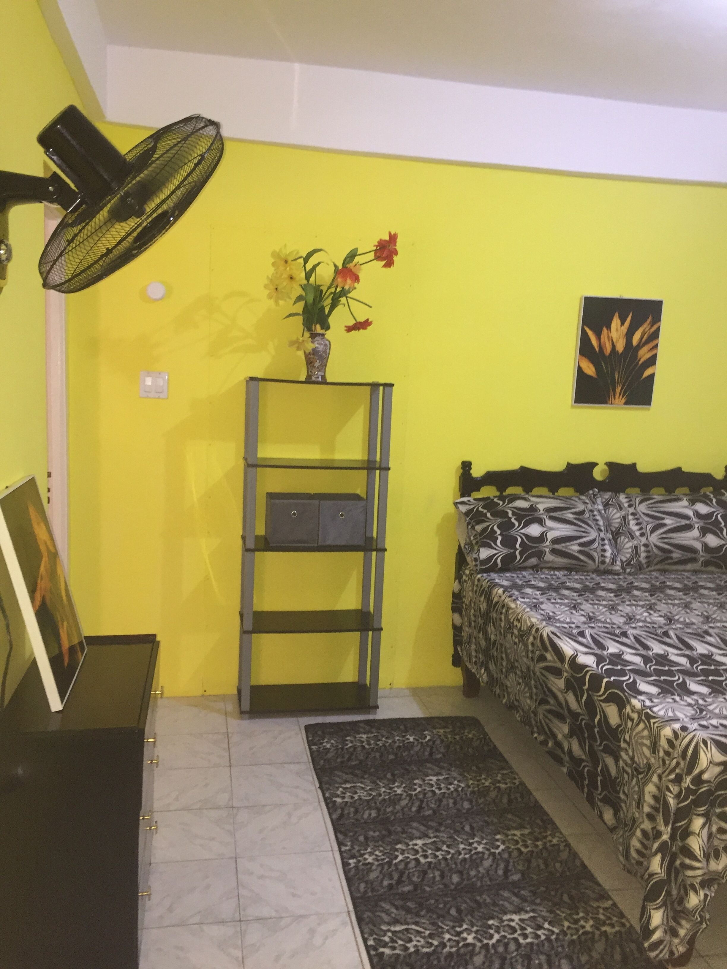 A room with a bed, a standing fan, a shelf with a vase of flowers, and a framed picture on the wall.