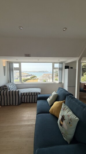 A spacious living room with a large window offering a view of the sea and hills. There is a blue sofa with cushions and a striped ottoman. - BellaVista 1B-Sleeps 4 in 2 Bedrooms (New Polzeath)