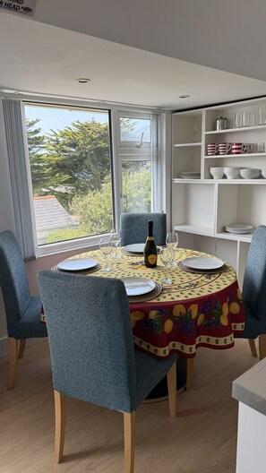 A dining area with a round table set for four, chairs, a bottle of Prosecco, and a view of trees through the window. - BellaVista 1B-Sleeps 4 in 2 Bedrooms (New Polzeath)