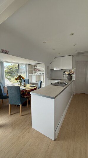 A modern kitchen with a central island, built-in shelving, and a dining area with a table and chairs. - BellaVista 1B-Sleeps 4 in 2 Bedrooms (New Polzeath)