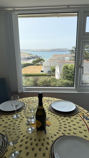 A dining table set for two with a bottle of wine, glasses, and plates, overlooking a coastal landscape. - BellaVista 1B-Sleeps 4 in 2 Bedrooms (New Polzeath)