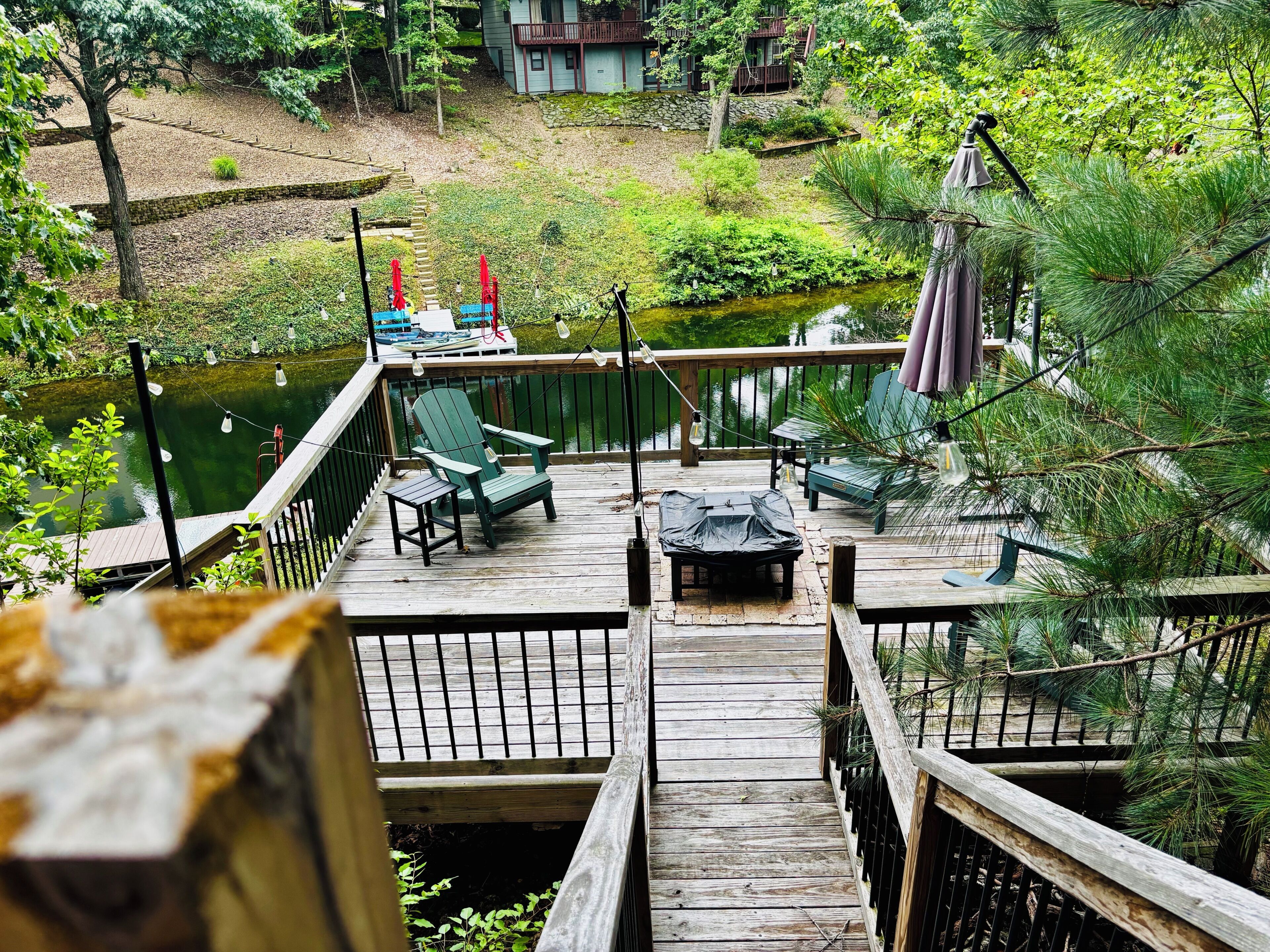 Une terrasse en bois, meublée pour l’extérieur, avec une petite table et une vue sur un étang et la verdure.