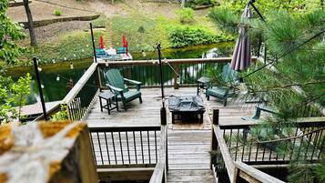 Une terrasse en bois, meublée pour l’extérieur, avec une petite table et une vue sur un étang et la verdure.