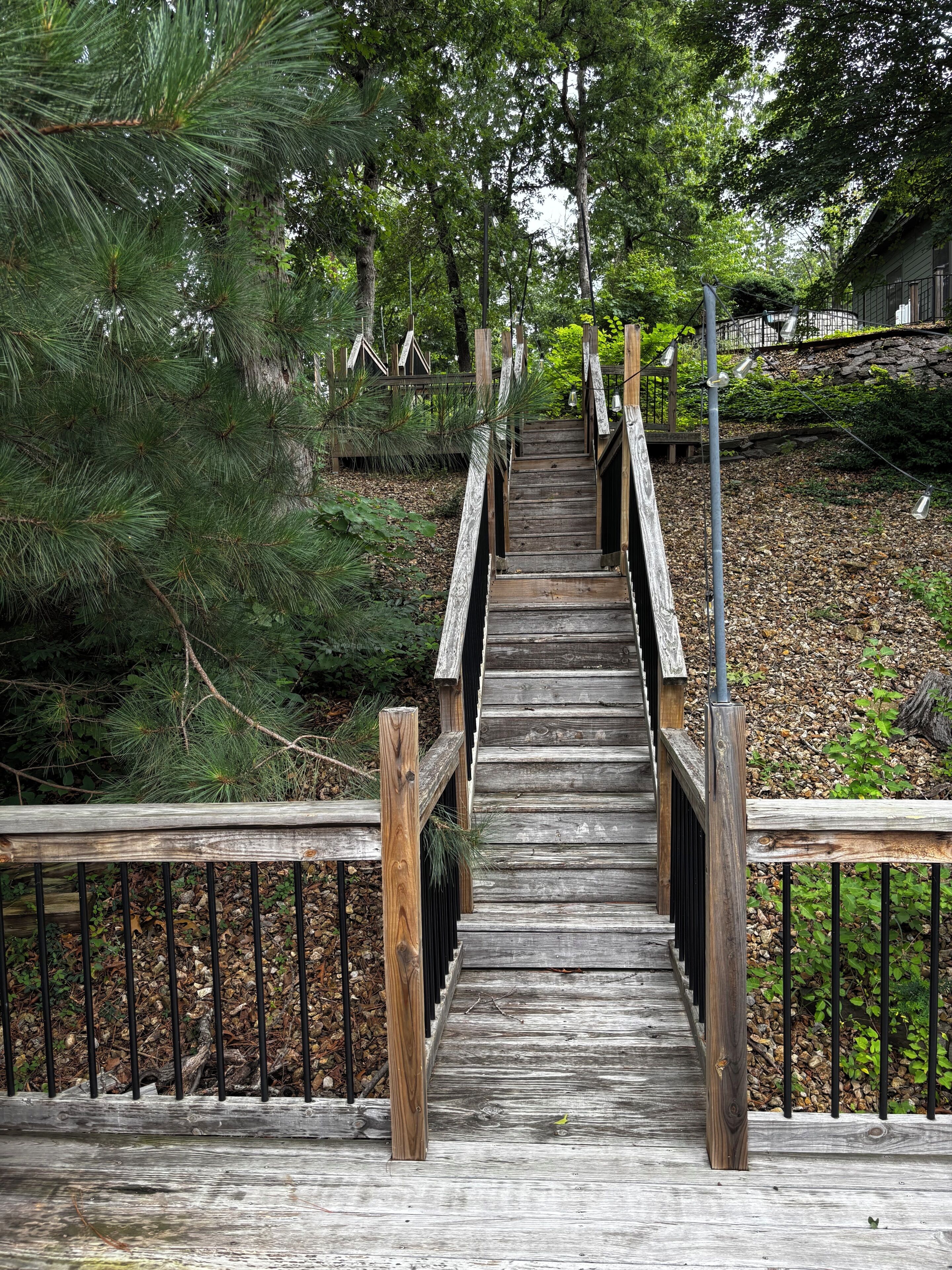 Eine Holztreppe führt zu einer Steinmauer, umgeben von Grünpflanzen und einem Metallgeländer.
