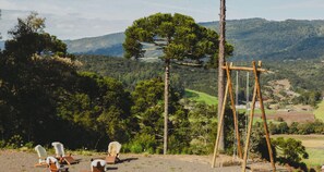 A rustic outdoor seating area with wooden chairs and a swing set, overlooking a lush valley with trees and distant hills.
