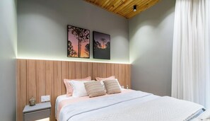 A bedroom with a wooden headboard, a bed with multiple pillows, a nightstand with a lamp, and two wall-mounted pictures.