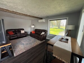 Ett vardagsrum med en mörkbrun lädersoffa, en mönstrad matta, ett matbord med barstolar och ett stort fönster med persienner.