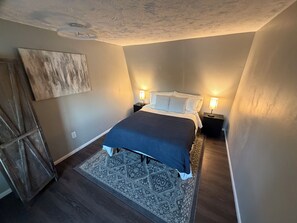 A bedroom with a bed, two bedside tables with lamps, a patterned rug, and a ceiling with a textured design.