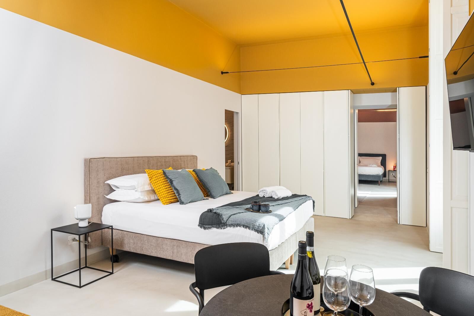 A modern hotel room with a bed, bedside table, and a dining area with chairs and a table.