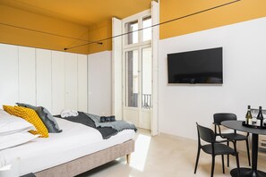 A bedroom with a bed, a TV, a dining table with chairs, and a balcony. - Loggiato Verga 18th Cent   Suite Carrara Bed&Bros (SIRACUSA)