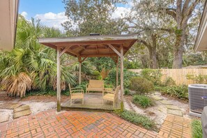A wooden pergola with a brick patio, surrounded by lush greenery and a wooden fence. - Tabby and Teal Cottage SSI (St. Simons Island)