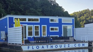 Ein blau-weißes Haus auf dem Wasser mit einer Terrasse, Sitzgelegenheiten im Freien und einer sichtbaren Webadresse.
