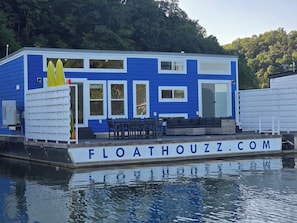 Ein blau-weißes Haus auf dem Wasser mit einer Terrasse, Sitzgelegenheiten im Freien und einer sichtbaren Webadresse.