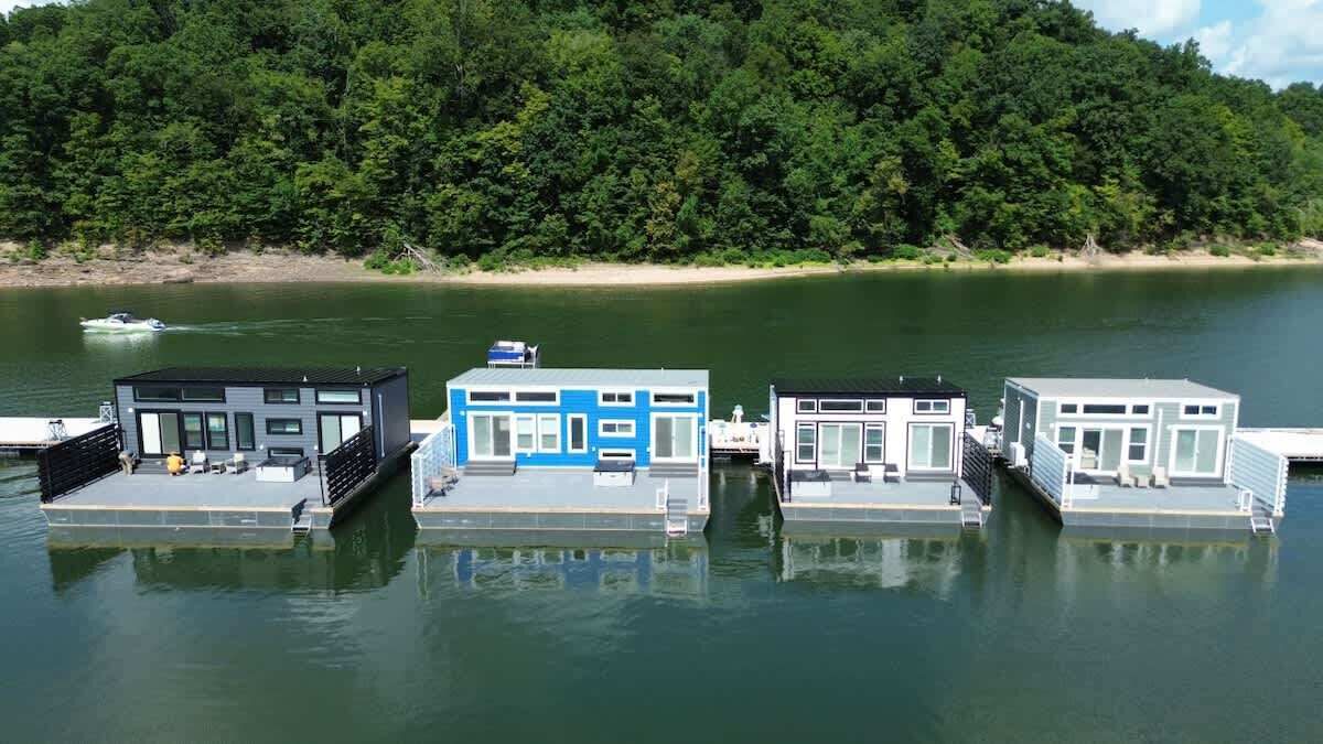 Trois maisons flottantes sur un lac paisible, bordé d’une rive boisée.