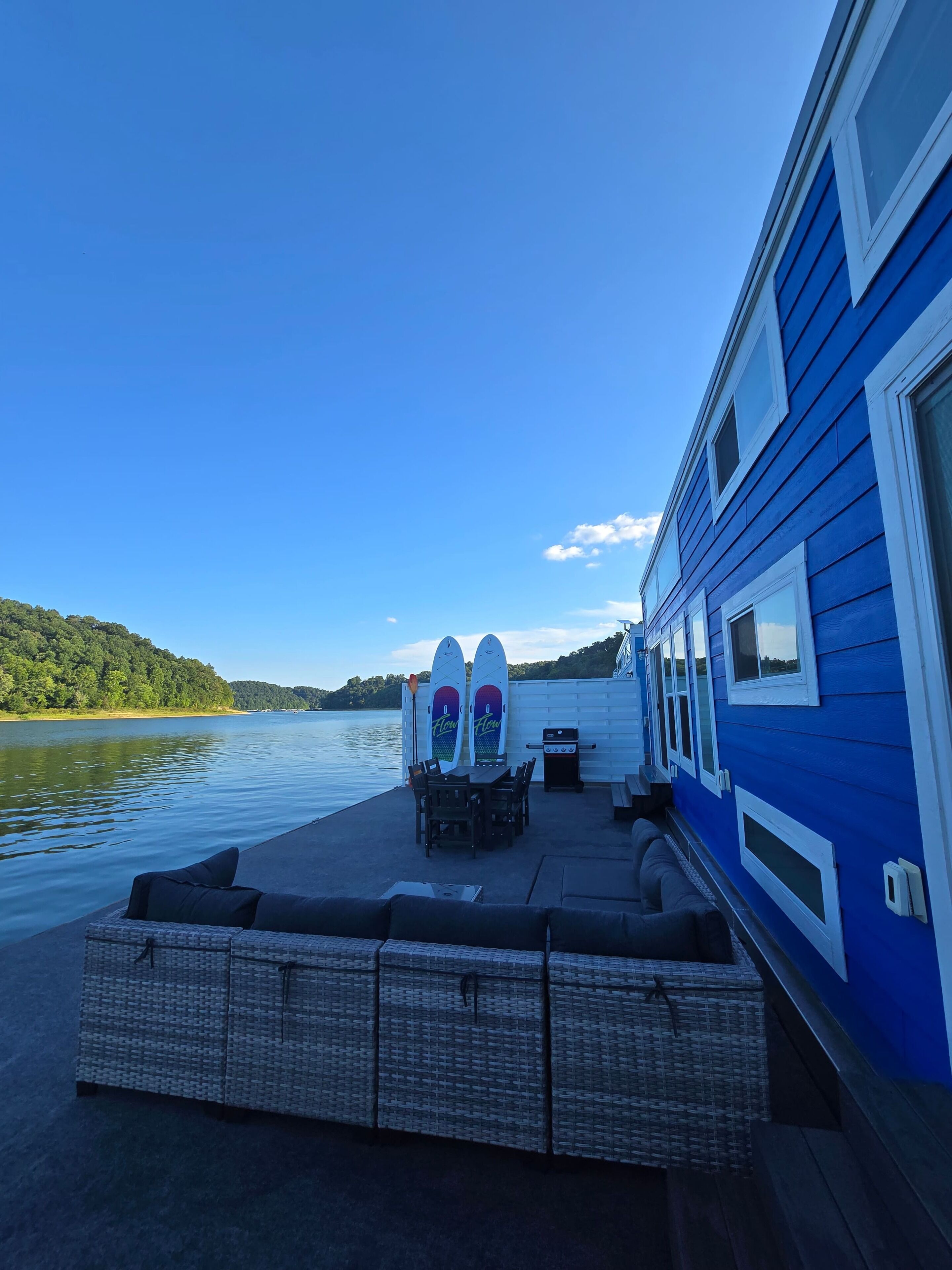 Une terrasse au bord de l’eau, avec une table et des chaises pour les repas, un barbecue et deux planches de surf appuyées contre le mur.