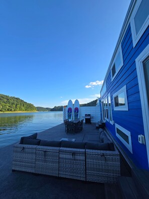 Une terrasse au bord de l’eau, avec une table et des chaises pour les repas, un barbecue et deux planches de surf appuyées contre le mur.