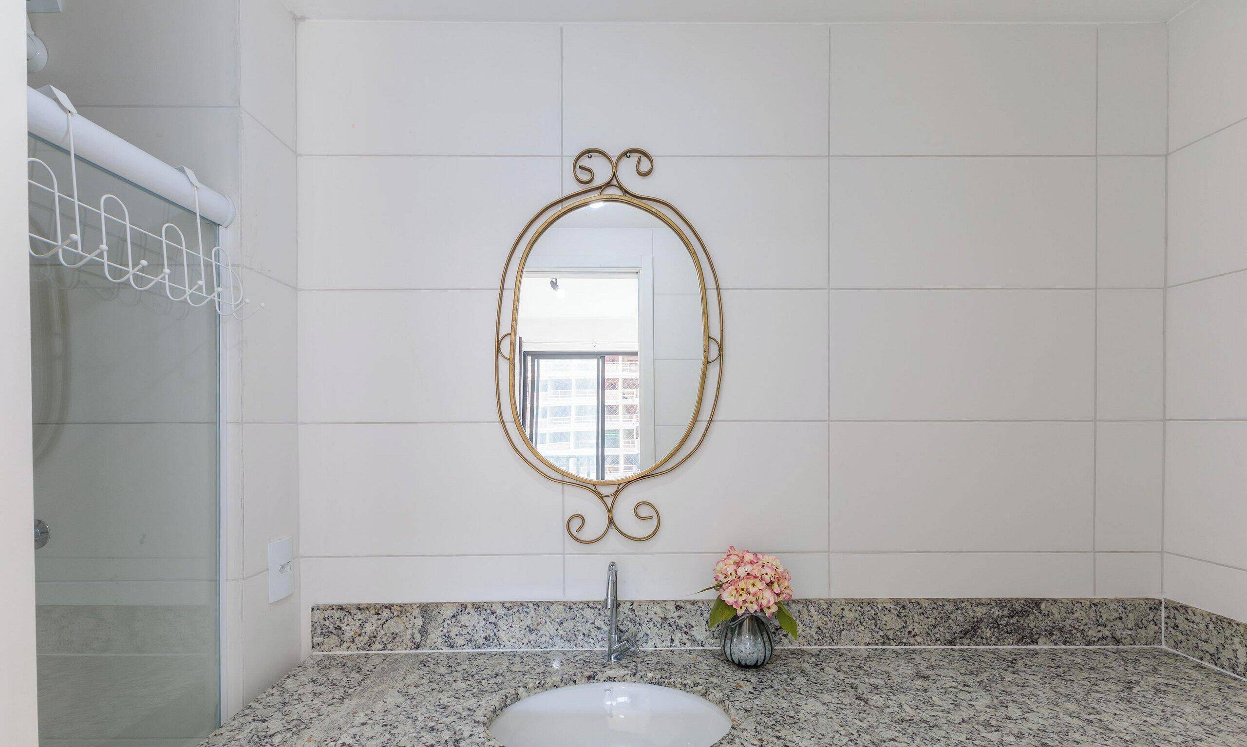 Baño con pared de azulejos blancos, un espejo independiente con marco dorado, encimera de granito, lavamanos y un florero con flores.