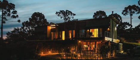 A modern two-story house with a balcony and a lit pool area at dusk.