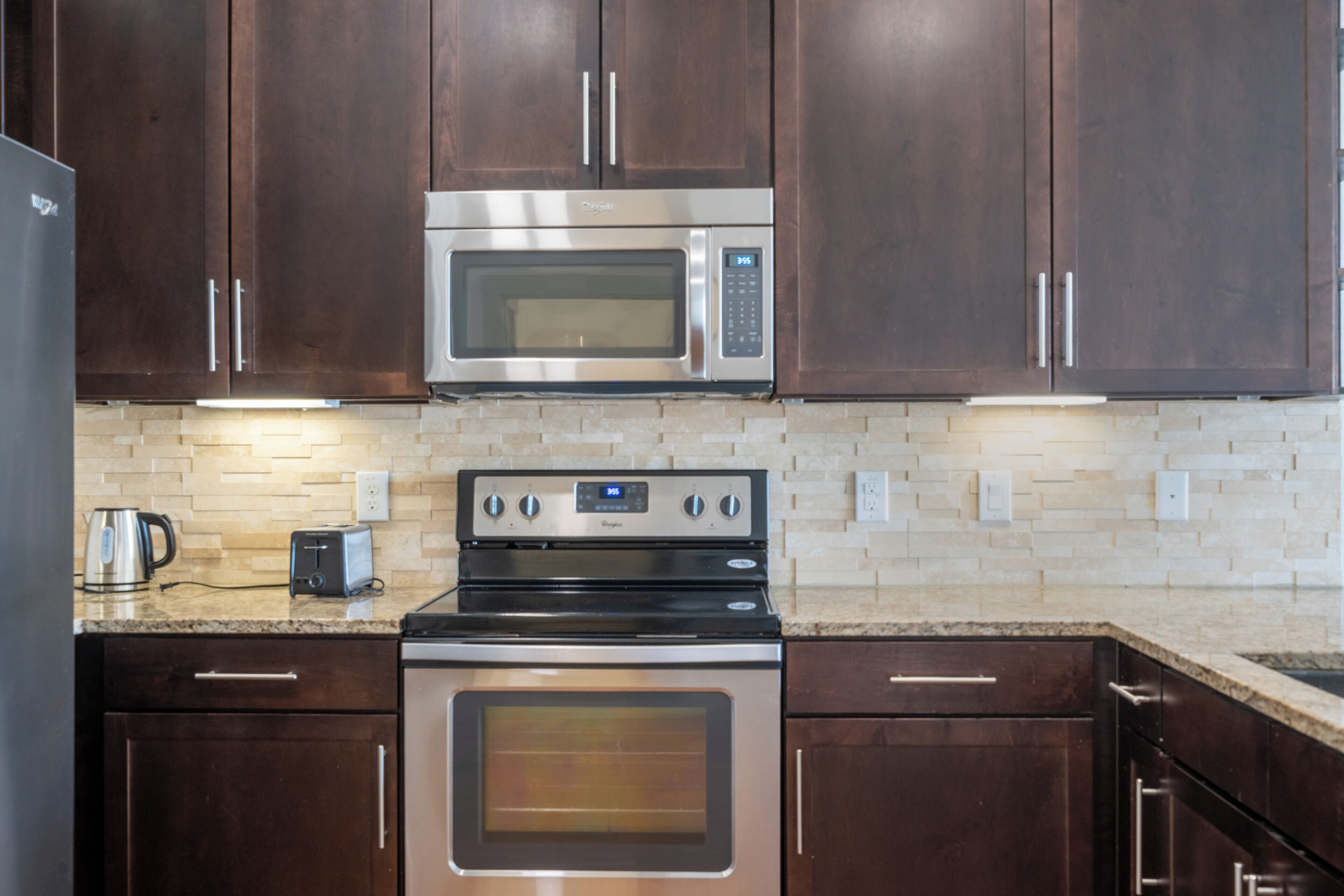 A kitchen with dark wooden cabinets, a stainless steel microwave, a built-in oven, a toaster, and a tiled backsplash.