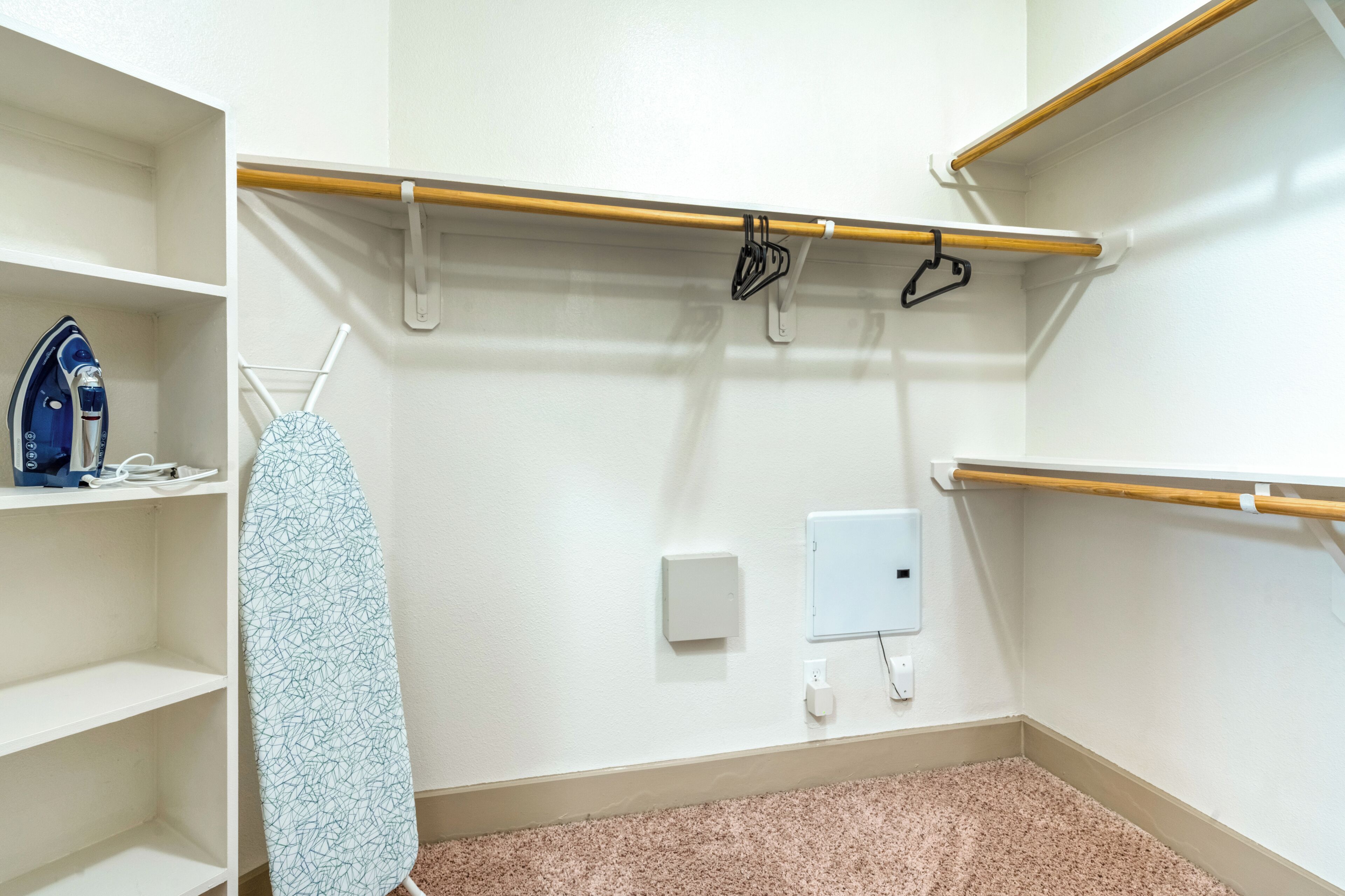 A hotel room with a white wall, a wooden shelf, a hanging rack with clothes, a blue iron, and a white electrical outlet.