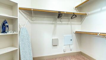 A hotel room with a white wall, a wooden shelf, a hanging rack with clothes, a blue iron, and a white electrical outlet.