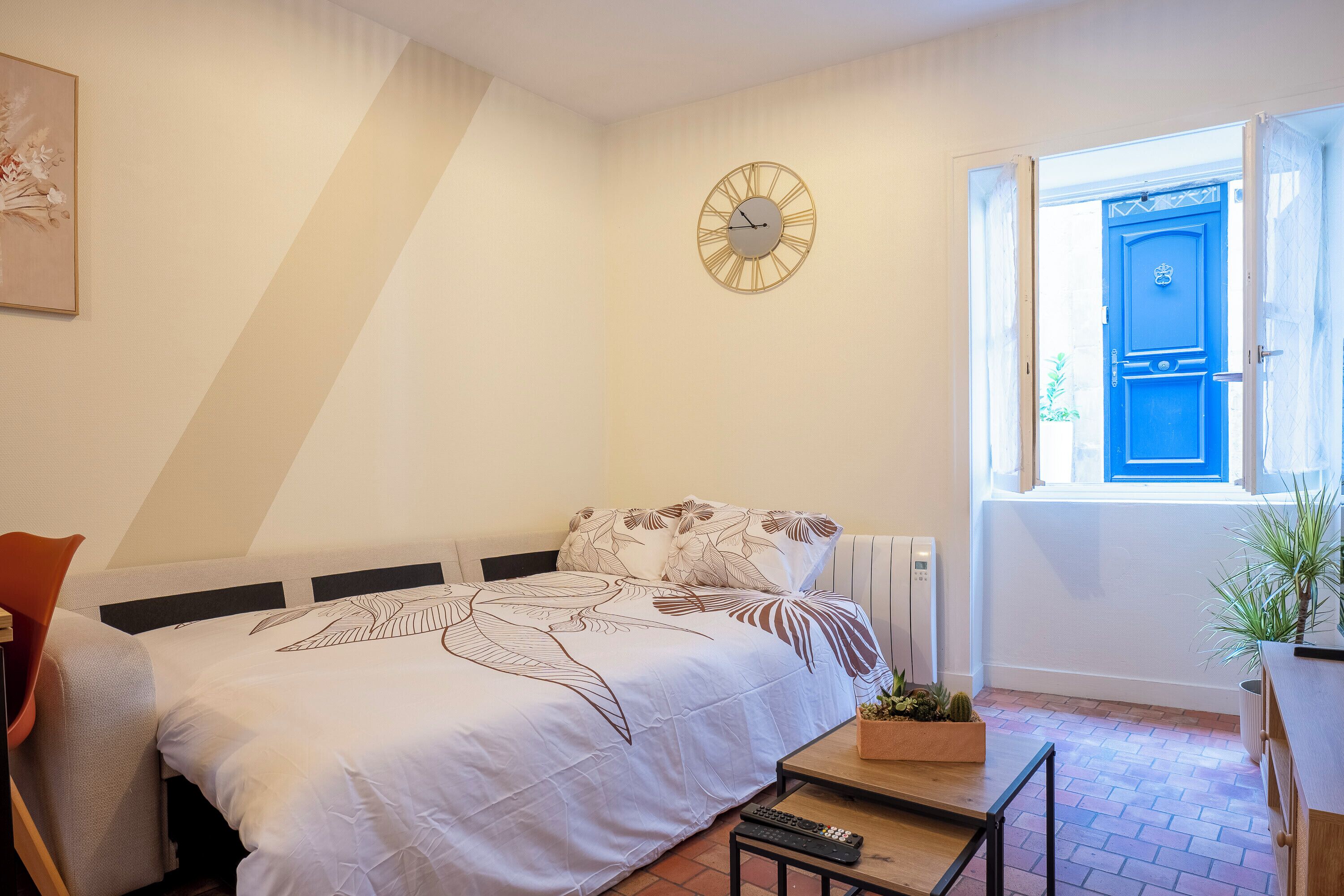 A bedroom with a bed, a bedside table, a clock, a window with a blue door, and a potted plant.