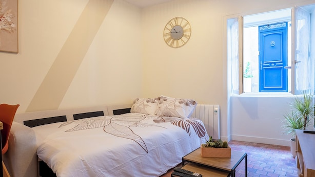 Ein Schlafzimmer mit einem Bett, einem Nachttisch, einer Uhr, einem Fenster mit blauer Tür und einer Topfpflanze.