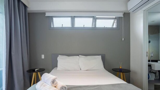 Quarto de hotel moderno com uma cama, duas mesas de cabeceira, uma janela e um banheiro visível através de uma porta aberta.
