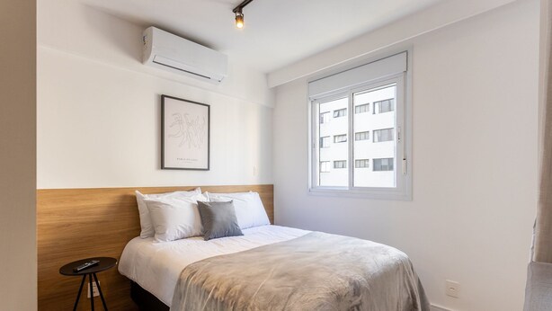 A bedroom with a bed, a small side table, a framed picture, an air conditioning unit, and a window with a view of buildings. - Complete Studio 500m From the Subway Cpm1110 (São Paulo)